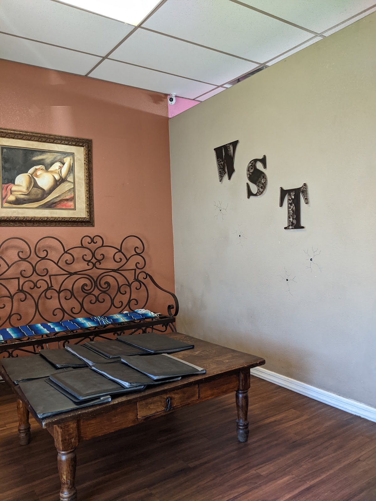 Waiting area with a wrought iron bench, striped serape blanket and gold framed painting on a terracotta wall.