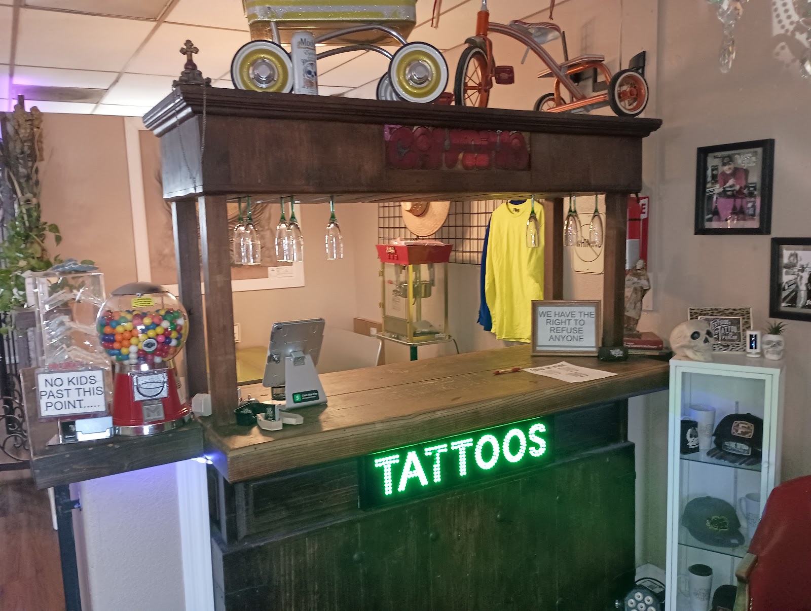 The front counter at Westside Tattoo: a wooden cantina style bar with a red gumball machine, vintage pedal cars on top, shop shirts and hats, and a green TATTOOS sign.