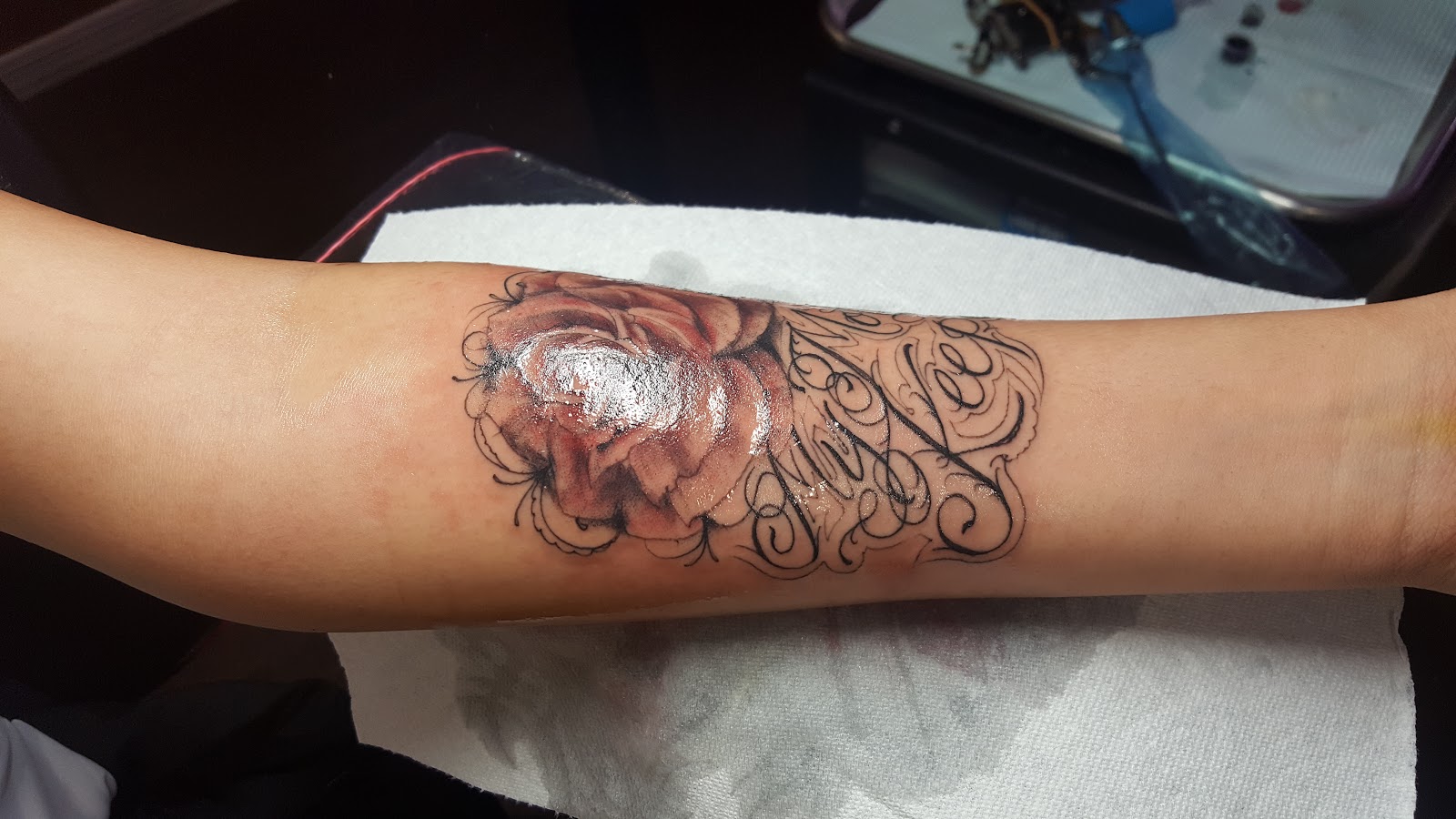 Black and grey rose with fine script lettering on a forearm, freshly done.